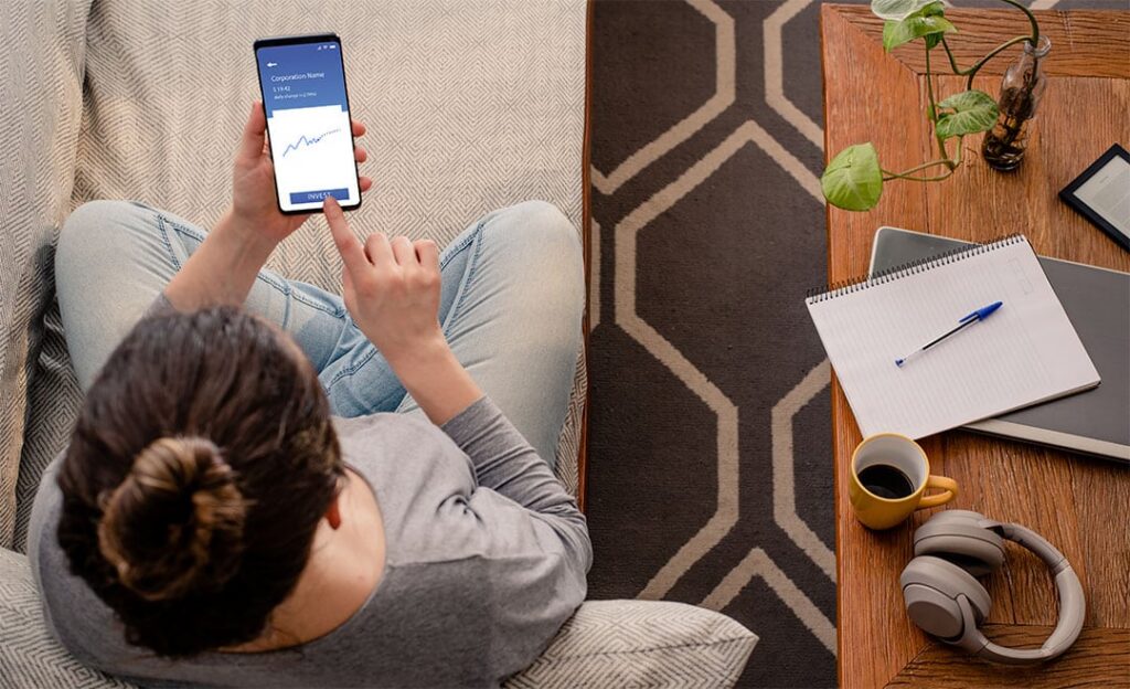 young investor sitting at home looking at her mobile investment app and hovering over the invest button on screen